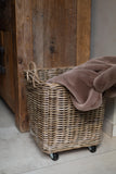 A square wicker storage basket with black caster wheels, filled with a dusty taupe throw blanket, set against a rustic wooden cabinet and light tile floor.