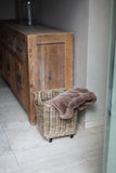 A natural wicker laundry hamper with black caster wheels, filled with a dusty taupe throw blanket, positioned next to a rustic wooden credenza in a neutral-toned room.