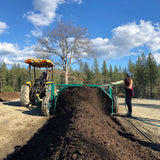 Tractor plowing a field with a person holding a hose, surrounded by trees and blue sky. Catalyst Craft Compost