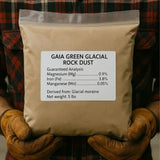 Bag of Gaia Green Glacial Rock Dust held by a person wearing gloves, with a plaid background.