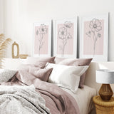 Neatly made bed with pink and white bedding, framed Pink Wall Art Set Of 3 Botanical Modern Decor on the wall, and a lamp on a side table.