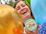 Woman holding a container of 'Rosemary Mint' body butter with balloons in the background