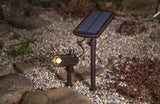 Solar-powered outdoor light fixture on a gravel surface with plants in the background