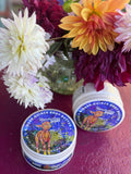 Two containers of 'Ginger Quince' body butter with colorful labels on a pink surface with flowers in the background.