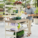Person working with plants and gardening tools on a multi-tiered shelving unit outdoors.