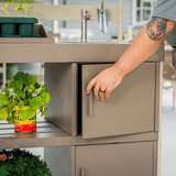 Person opening a cabinet door in a kitchen setting with plants and a sink visible.