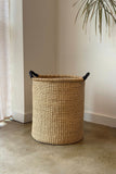 Woven basket with handles on a concrete floor next to a wall with a plant.