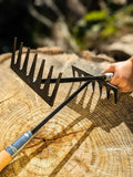 Two Hand Rake, Hand Forged Garden Tools with wooden and metal handles resting on a cut tree stump outdoors, with a person's hand holding one rake.