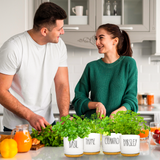 A man in a white t-shirt and a woman in a green sweater stand in a bright kitchen, smiling at each other. In the foreground, four white planters labeled Basil, Thyme, Cilantro, and Parsley sit on a counter filled with fresh green herbs.