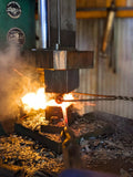 A blacksmith's forge in action, showing a glowing red-hot piece of metal being hammered by a large press with bright orange and yellow flames, set against a rustic workshop background.
