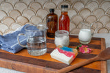 Wooden tray with glassware, bottles, and a bar of soap on a tiled wall background
