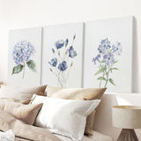 Three floral paintings on a wall above a bed with pillows and a lamp.