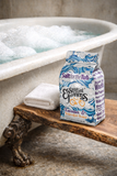 Bag of 'Salt for the Tub' by Essential Elements on a wooden shelf next to a bathtub with bubbles.
