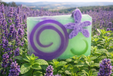 Bar of LAVENDER MINT SOAP SLICE 5.5 oz. with a purple flower on top, surrounded by lavender plants.