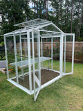Greenhouse with transparent walls and a metal frame on a grassy area with trees in the background. The Sago Greenhouse 5' x 8'