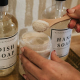 Hand holding a jar of soap with a wooden spoon, surrounded by dish soap and hand soap bottles. Agave Products