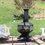 Black outdoor chiminea with flames on a patio with chairs and grass in the background