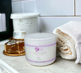 Jar of lavender body butter on a bathroom counter with a towel and jar of honey in the background.