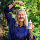 Woman in a purple shirt holding a bottle of lotion outdoors with greenery in the background