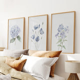 Three framed floral prints on a wall above a bed with pillows and a lamp.