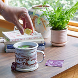Person adding a lemon wedge to a marbled mug of tea on a wooden table with books and a plant in the background Altered Herb Mug Grow Kits.