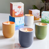 Four lit candles in colorful containers with boxes in the background on a white surface.