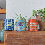 Three colorful sprout grow kits on a wooden surface with plants in the background.