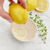 Hand squeezing a lemon over a bowl with lemons and herbs on a light background Peel, Grate & Prep Dish