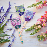 Four wooden seed pop sticks with colorful labels on a wooden surface surrounded by flowers. Seed Pops - Flowers