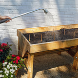Person watering a wooden raised garden bed with a hose against a light gray brick wall.