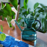 Blue glass carafe with handle on a wooden table with plants in the background