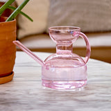 Pink Glass Watering Can on a marble surface with a blurred background