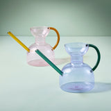Two glass watering cans with colorful handles on a light green background