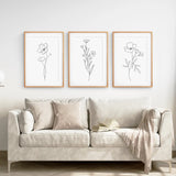 Beige sofa with decorative pillows and a blanket, featuring three framed botanical line art prints on a white wall.