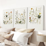 Three botanical wall art prints above a bed with pillows and a lamp.