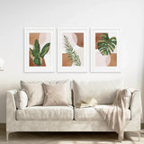 Beige sofa with decorative pillows and a blanket, featuring three framed 3 Piece Terracotta Boho Wall Art. Green Tropical Leaves on the wall.