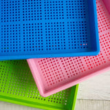 Three perforated plastic trays in blue, pink, and green on a wooden surface. 1020 Mesh Trays Shallow Extra Strength Colors -  1" Deep