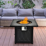 Outdoor fire pit table with flames on a wooden deck, surrounded by a gray sectional sofa and greenery.