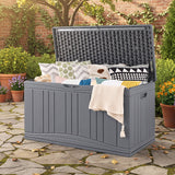 A large gray outdoor storage trunk with its lid open, styled with decorative pillows and a cream throw blanket in a garden patio setting.