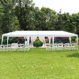 White outdoor canopy tent set up on a grassy area with tables and chairs, surrounded by trees.