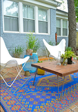 Outdoor patio with a colorful Turkish Blue rug, wooden table, and white chairs in front of a house.