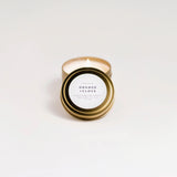 Candle in a gold tin labeled 'Orange + Clove' on a white background