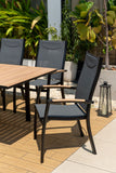 Outdoor dining set with wooden table and black chairs on a patio with plants in the background.