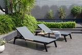 Two gray lounge chairs with a small table on a patio with plants and a wall in the background.