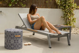 Woman relaxing on a sun lounger with a patterned ottoman and sandals nearby.