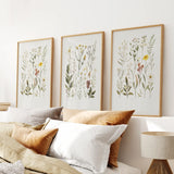 Three framed botanical prints above a bed with pillows and a lamp.