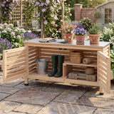 A wooden outdoor garden table with open slatted doors and shelves, shown in a garden setting. The table has a light natural wood finish, a gray stone-look top, and is styled with gardening tools, pots, plants, a bucket, and green boots.