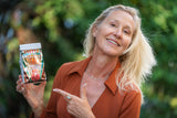 Woman holding a POMEGRANATE SUNRISE ARNICA BATH SALTS package with a blurred green background