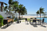 Outdoor patio area with dining table, chairs, and barbecue grill by a poolside with palm trees and beach in the background.