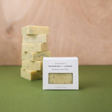 Bar of soap with packaging labeled 'Rosemary + Lemon' on a green surface with a wooden background.